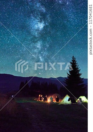 Group of friends hikers resting around campfire, surrounded by illuminated tents under stunning starry sky. Milky Way stretches above, creating magical atmosphere in peaceful mountain wilderness. 117041681