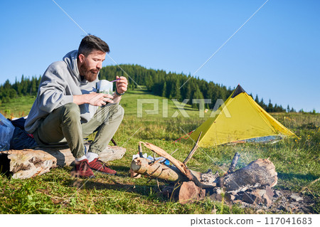 Man tourist eating from food pouch with green spoon, sits on log near campfire. Yellow tent on green hillside, surrounded by lush trees. Peacefulness of camping in beautiful natural environment. 117041683