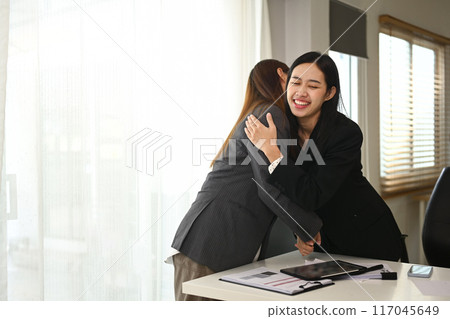 Happy cheerful businesswomen standing in office and embracing each other, celebrating good news Happy cheerful businesswomen standing in office and embracing each other, celebrating good news 117045649