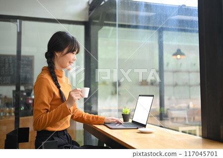Pretty young Asian woman in warm sweater drinking coffee and using laptop in bright cafe 117045701