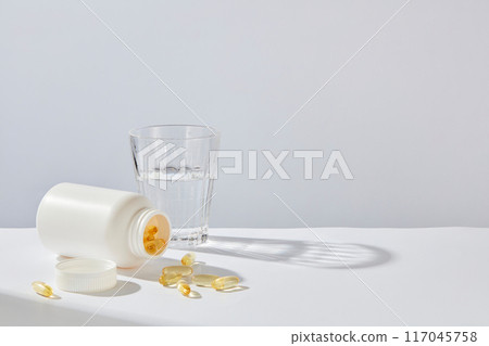 Front angle shot photography for nutrient medication advertisement on white background. A unlabeled jar lay on the table, next to a glass of water and some flat lay yellow capsules Front angle shot photography for nutrient medication advertisement on white background. A unlabeled jar lay on the table, next to a glass of water and some flat lay yellow capsules 117045758