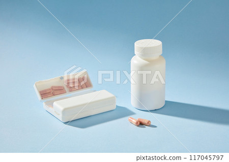 The medicine theme photograph with simple advertising space, from frontal shot. An empty label drug bottle in white displayed next to white pill box, which contains some light purple pills 117045797