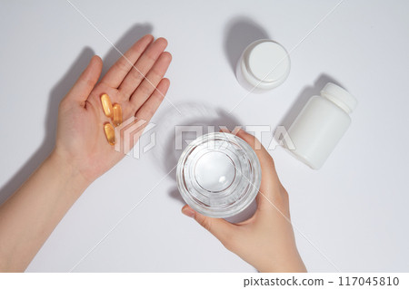 Photo scene of the preparation for medicine drinking on white background from top angle. Female's hand with some yellow pills in palm, the other one holding a water glass, next to some unlabeled jars 117045810