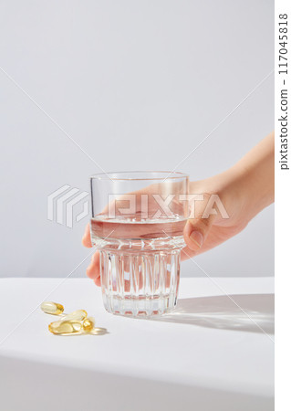 Minimalism photograph of a hand holding a water glass, which placed in the center of photo, next to some yellow vitamin tablets flat lay on white table top. Front view, space to design with sunlight Minimalism photograph of a hand holding a water glass, which placed in the center of photo, next to some yellow vitamin tablets flat lay on white table top. Front view, space to design with sunlight 117045818