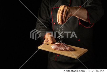 Chef sprinkles salt on meat steak with hand before grilling. Hotel or restaurant service concept on black background with place for advertising. Cooking meat in kitchen Chef sprinkles salt on meat steak with hand before grilling. Hotel or restaurant service concept on black background with place for advertising. Cooking meat in kitchen 117046454