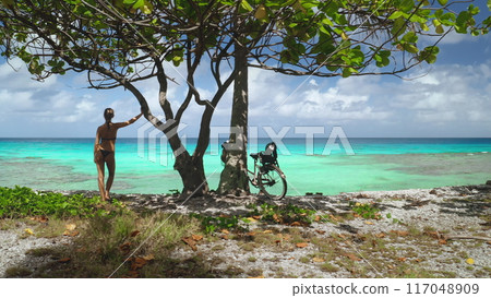 Tourist woman in swimsuit stands by tree on a tropical beach in French Polynesia. Turquoise ocean water, clear blue sky in warm, sunny day, tranquil and peaceful atmosphere. Summer holiday vacation Tourist woman in swimsuit stands by tree on a tropical beach in French Polynesia. Turquoise ocean water, clear blue sky in warm, sunny day, tranquil and peaceful atmosphere. Summer holiday vacation 117048909