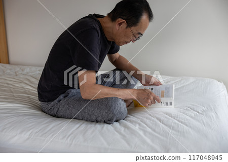 A man sitting cross-legged on the bed and reading a book A man sitting cross-legged on the bed and reading a book 117048945