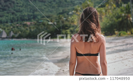 Woman is walking on a tropical beach, enjoying the warm sun and gentle breeze as she takes in the beautiful ocean view Woman is walking on a tropical beach, enjoying the warm sun and gentle breeze as she takes in the beautiful ocean view 117048950
