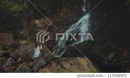 Woman sitting big stone, take picture on mobile phone of waterfall cascade rocky cliff. Female in white shirt relax enjoy beautiful wild nature landscape exotic rainforest. Travel tropical Bali island Woman sitting big stone, take picture on mobile phone of waterfall cascade rocky cliff. Female in white shirt relax enjoy beautiful wild nature landscape exotic rainforest. Travel tropical Bali island 117048955