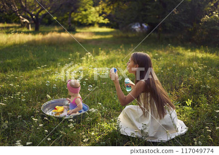a baby in a metal cup in a summer garden plays with rubber ducklings, a teenage sister makes soap bubbles 117049774