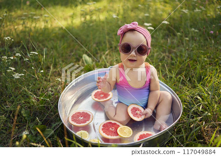 baby wearing sunglasses in a metal bath in a summer garden plays with grapefruit and lemon slices 117049884