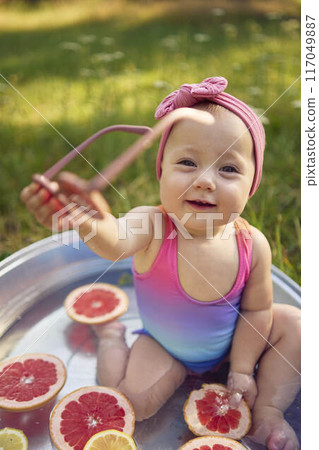 baby wearing sunglasses in a metal bath in a summer garden plays with grapefruit and lemon slices 117049887
