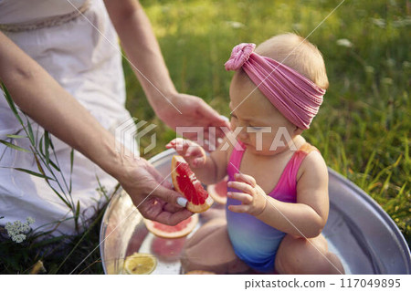 baby in metal bath in summer garden watching mother and teenage sister eat grapefruit baby in metal bath in summer garden watching mother and teenage sister eat grapefruit 117049895