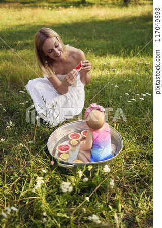 baby in metal bath in summer garden watching mother and teenage sister eat grapefruit 117049898