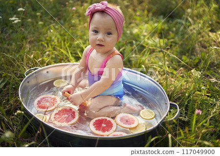 a baby in a metal bath in a summer garden plays with grapefruit and lemon slices 117049900