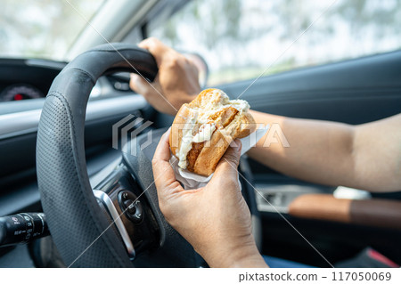 Asian lady holding hamburger to eat in car, dangerous and risk an accident. Asian lady holding hamburger to eat in car, dangerous and risk an accident. 117050069