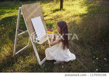 pre-teen girl in a summer dress paints a picture in the garden on an easel pre-teen girl in a summer dress paints a picture in the garden on an easel 117050106
