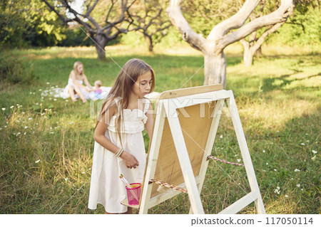 mother and baby sister watch as  pre-teen girl in a summer dress paints a picture in the garden on an easel 117050114