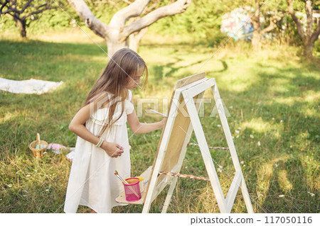 pre-teen girl in a summer dress paints a picture in the garden on an easel pre-teen girl in a summer dress paints a picture in the garden on an easel 117050116