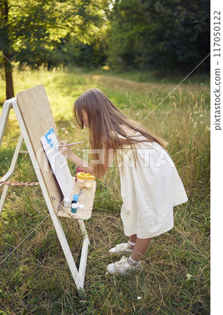 pre-teen girl in a summer dress paints a picture in the garden on an easel pre-teen girl in a summer dress paints a picture in the garden on an easel 117050122