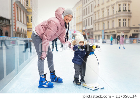 Young adult attractive beautiful caucasian woman enjoy having fun with little toddler son at outdoor skating rink while travel in old european city in winter. Mom and child holiday sport acitivities Young adult attractive beautiful caucasian woman enjoy having fun with little toddler son at outdoor skating rink while travel in old european city in winter. Mom and child holiday sport acitivities 117050134