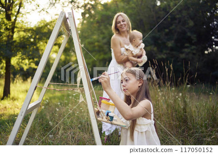 mother and baby sister watch as pre-teen girl in a summer dress paints a picture in the garden on an easel mother and baby sister watch as pre-teen girl in a summer dress paints a picture in the garden on an easel 117050152