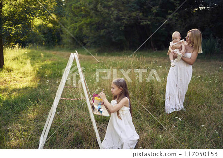 mother and baby sister watch as  pre-teen girl in a summer dress paints a picture in the garden on an easel 117050153