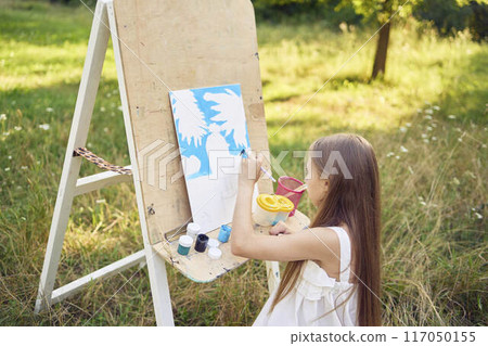 a pre-teen girl in a summer dress paints a picture in the garden on an easel a pre-teen girl in a summer dress paints a picture in the garden on an easel 117050155