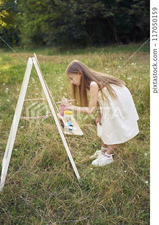 a pre-teen girl in a summer dress paints a picture in the garden on an easel 117050159
