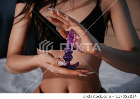 silicone butt anal plug in hands of a woman with a sexy body and breasts in bra sitting on bed in bedroom. Female sex toys for anal sex and orgasm 117052814