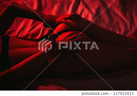 submissive woman girl with a sexy body and ass in panties with handcuffed hands lying on bed in bedroom. BDSM sex with submission and domination with red neon light 117052815