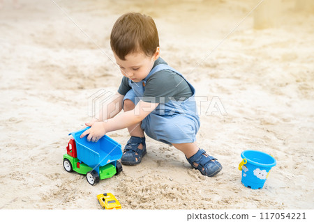 Little cute toddler boy 2.5 years old plays in the sandbox on a sunny summer day. Outdoor creative activities for kids. Toy car and shovel. 117054221