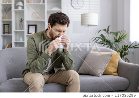 Man sneezing into tissue while sitting on the couch. Indoor scene with man having cold or allergies, showing discomfort and illness. 117056299