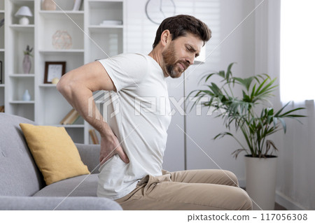 Man sitting on couch holding lower back, experiencing back pain, discomfort or injury. Concept of health issues, chronic pain, posture problems, or muscle strain. Man sitting on couch holding lower back, experiencing back pain, discomfort or injury. Concept of health issues, chronic pain, posture problems, or muscle strain. 117056308