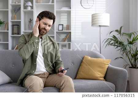 Man sitting on couch experiencing ear pain while holding smartphone in hand. He looks uncomfortable and in pain. Home setting with modern decor visible in background. 117056315