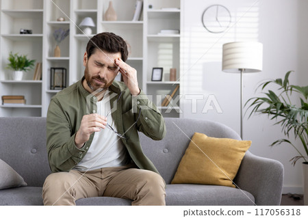 Man experiencing headache sitting on couch at home, touching temples and holding glasses. Concept of stress, pain, fatigue in casual setting with modern furniture. 117056318