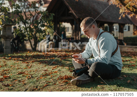 Professional Photographer Taking Picture Of Beautiful Autumn Park. Man Professional Photographer Sit With Camera And With Smartphone In Autumn Park. Retouched, Vibrant Colors, Brownish Tones. 117057505