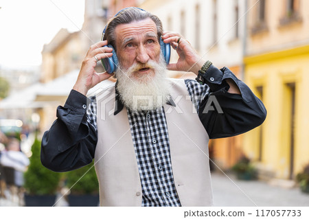 Happy senior old man in wireless headphones choosing, listening music dancing outdoors city street 117057733