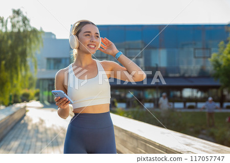Happy young woman jogger listening to music via headphones and smartphone walking in city park Happy young woman jogger listening to music via headphones and smartphone walking in city park 117057747