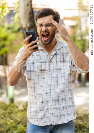 Angry mad Hispanic young man arguing while talking on smartphone irritated shout quarrel outdoors Angry mad Hispanic young man arguing while talking on smartphone irritated shout quarrel outdoors 117057752