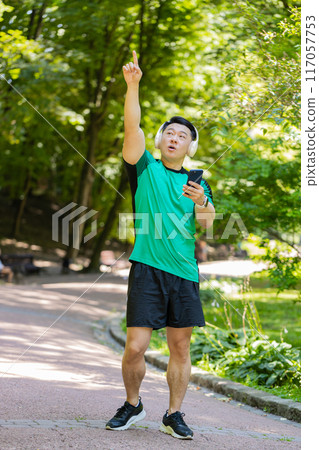 Athletic fitness Asian sport runner man walking, wearing headphones listening music from smartphone Athletic fitness Asian sport runner man walking, wearing headphones listening music from smartphone 117057753