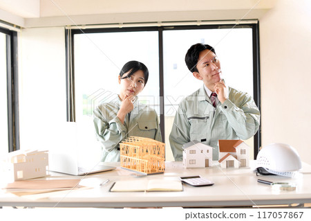 Asian man and woman in work clothes worried in front of a model of a house Asian man and woman in work clothes worried in front of a model of a house 117057867