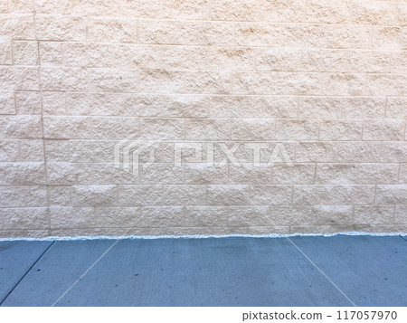 A section of a beige brick wall with a clean, smooth concrete sidewalk in the foreground. The wall textured surface and uniform brick pattern provide a neutral backdrop, while the sidewalk adds a 117057970