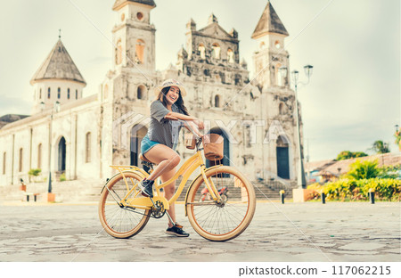 Happy girl in hat riding a bicycle at sunset. Tourism and travel concept. Lifestyle of a happy girl in hat on bicycle on the street. Granada, Nicaragua 117062215