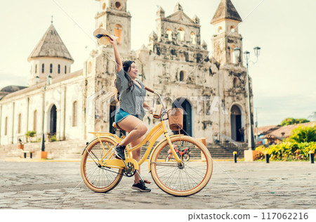 Lifestyle of a happy girl riding a bicycle holding her hat on the street. Happy young woman in hat riding a bicycle on the street. Granada, Nicaragua. Tourism concept 117062216
