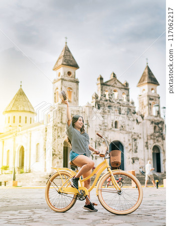 Happy young woman in hat riding a bicycle on the street. Granada, Nicaragua. Lifestyle of a happy girl riding a bicycle holding her hat on the street. Tourism concept Happy young woman in hat riding a bicycle on the street. Granada, Nicaragua. Lifestyle of a happy girl riding a bicycle holding her hat on the street. Tourism concept 117062217