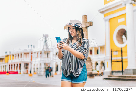 Female tourist texting phone in the square of Granada. Smiling young tourist in hat using cell phone on the street 117062274