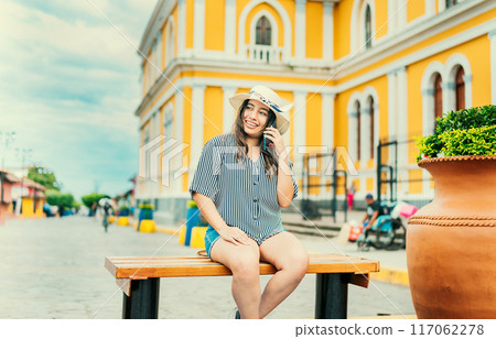 Smiling tourist sitting on a bench talking on cell phone. Happy travel woman sitting calling on the phone in a tourist square 117062278