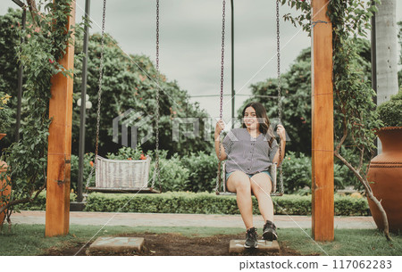 Portrait of smiling girl sitting on a swing in a garden. Happy young woman sitting on a swing in a beautiful garden. 117062283