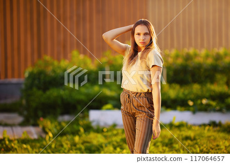 Portrait of young Caucasian woman standing sideways, half-turned, knee-length horizontal photo, during golden hour in countryside on summer evening. Portrait of young Caucasian woman standing sideways, half-turned, knee-length horizontal photo, during golden hour in countryside on summer evening. 117064657